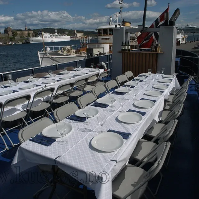 Dekket selskapsbåt med hvite duker, tallerkener og glass, klart for gjester ved kai i havneområde.