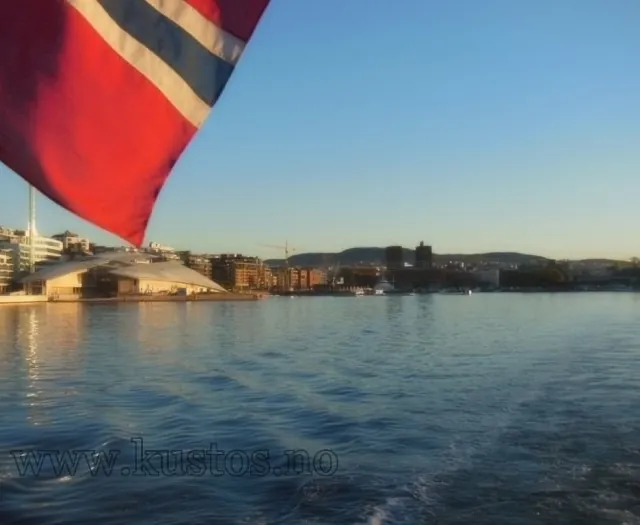 Utsikt fra båt over havn og by i solnedgang med norsk flagg i forgrunnen.