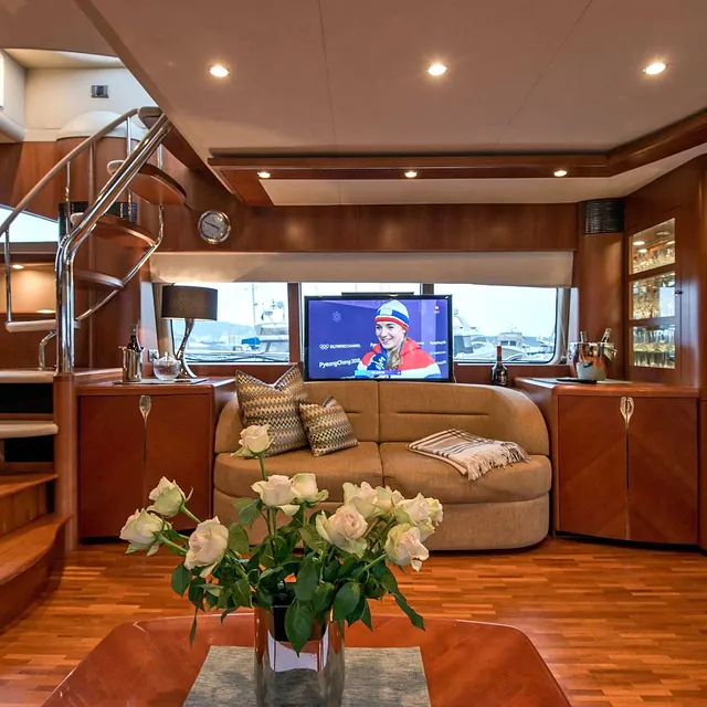 TV-stue på yacht med buet sofa, blomster på bordet og TV foran vinduer med havneutsikt