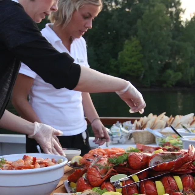 To kvinner serverer sjømatbuffé med krabbe, reker og hummer på utendørs dekk.