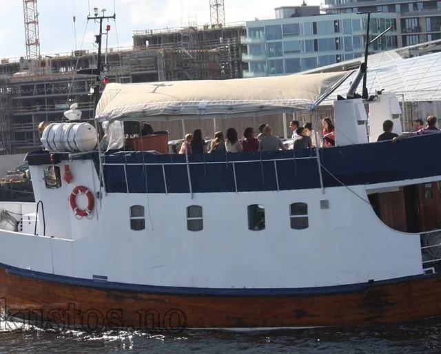 Mindre trebåt med hvit overbygning og passasjerer på dekk, seilende forbi byggeplass i havneområde.