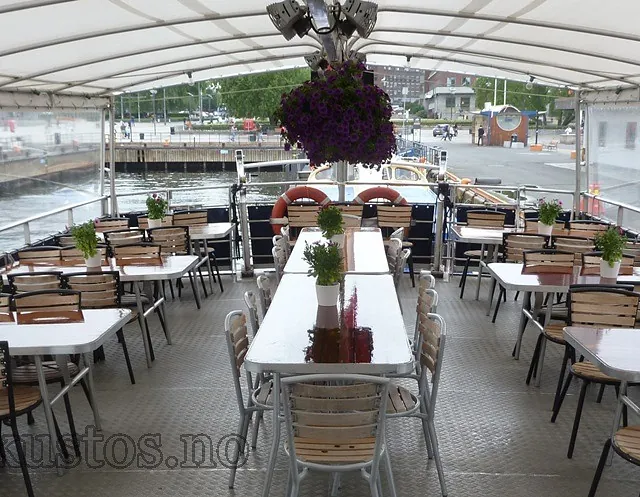 Overdekket spiseområde på båt med bord, stoler og grønne planter i potter.