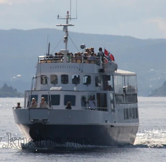 Fjordclipper i fart med mange passasjerer ombord og norsk flagg bak.